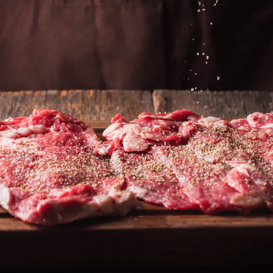 Raw beef steaks spread on a wooden board with salt being sprinkled over them