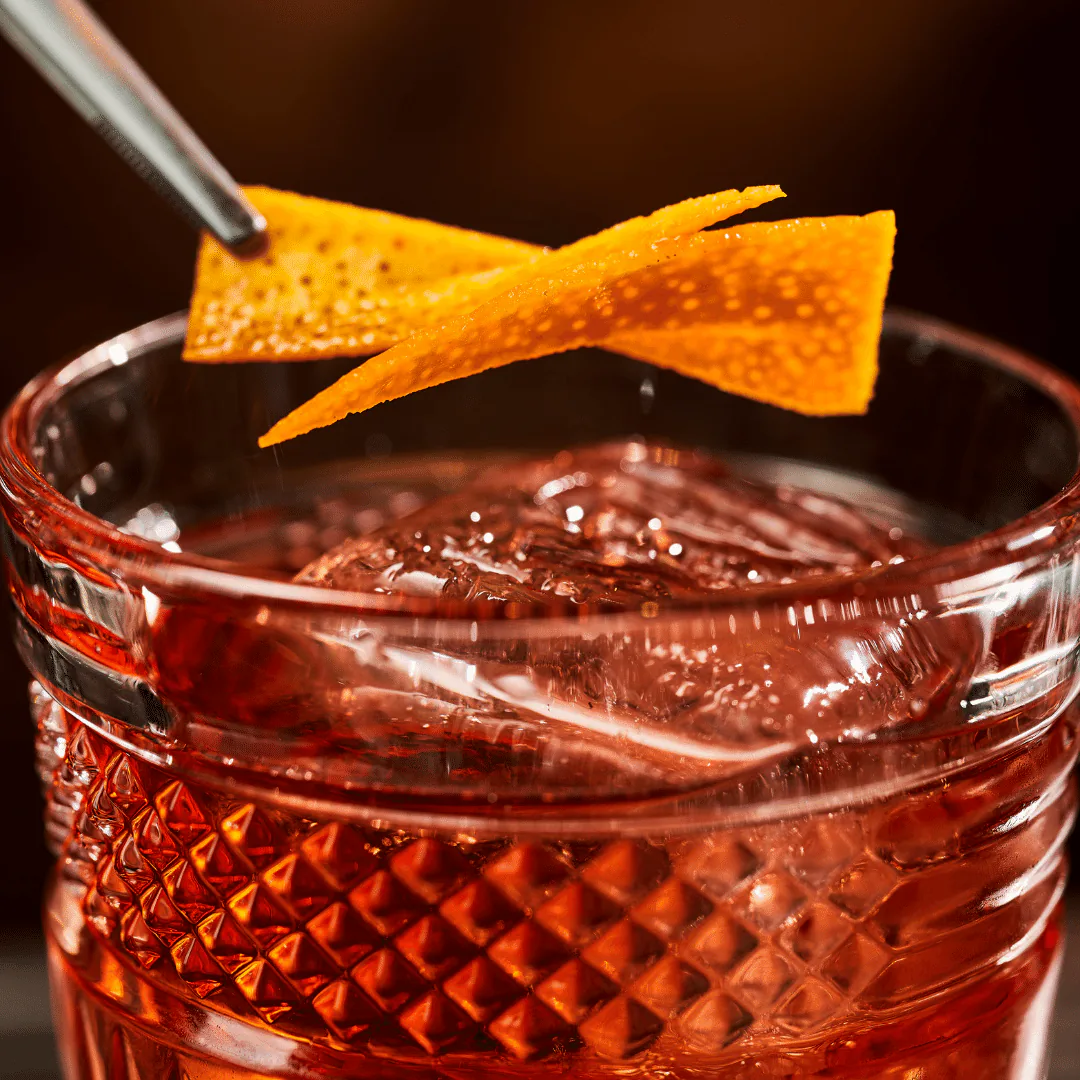 Close-up of a red cocktail in a cut-crystal glass with a curled orange peel twist and a bar spoon