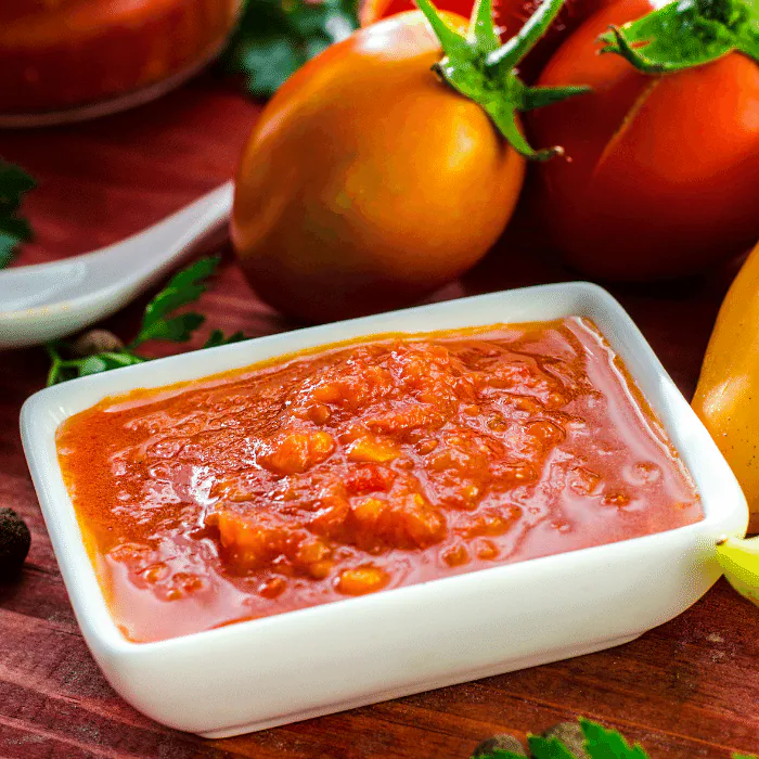 Chunky red pepper hot sauce in a white dish with fresh tomatoes and a yellow chili behind