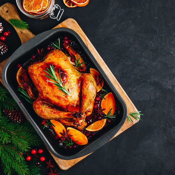 Whole roast chicken in a pan with cranberries, orange slices and rosemary, framed by pine branches