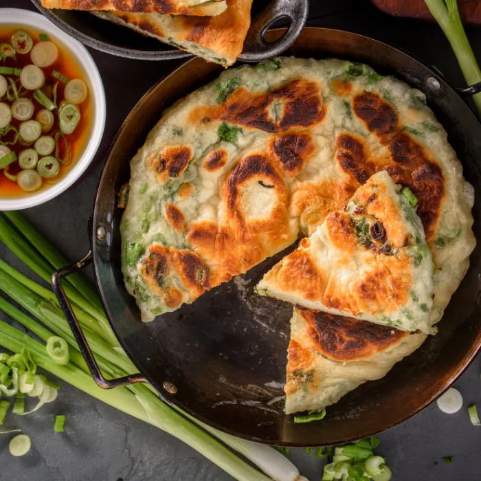 Golden scallion pancake sliced into wedges in a steel pan with dipping sauce and fresh scallions