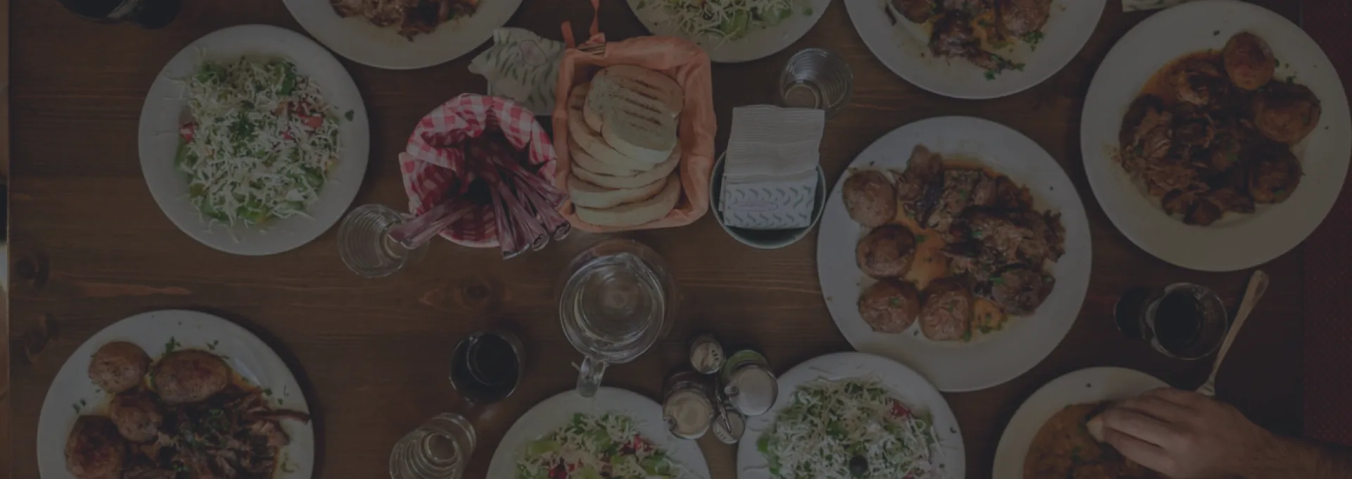 Overhead view of a wooden table spread with shared plates of roast potatoes, salads and flatbread