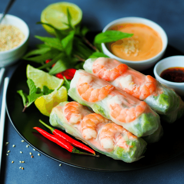 Rice paper summer rolls filled with shrimp and greens on a dark plate with peanut sauce, chilies and lime