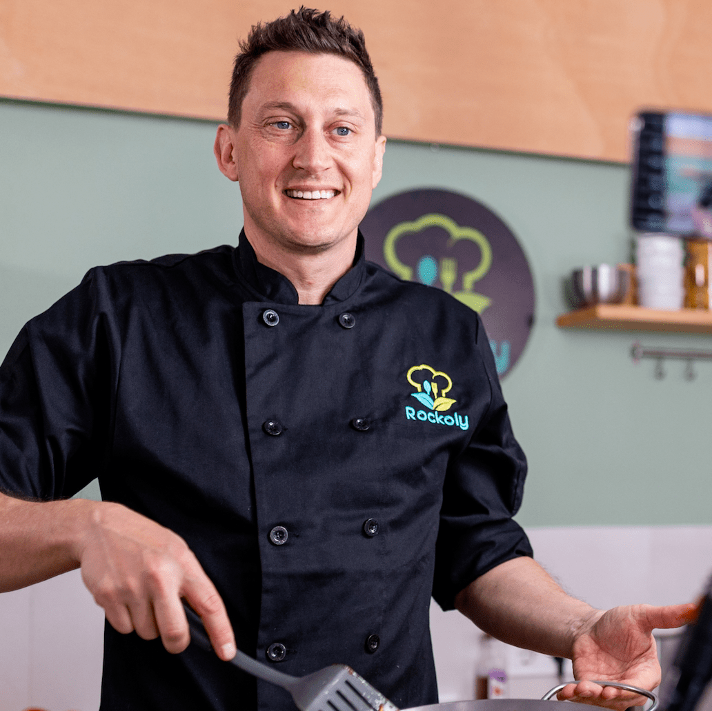 Smiling chef in a black Rockoly chef coat gesturing while leading a class
