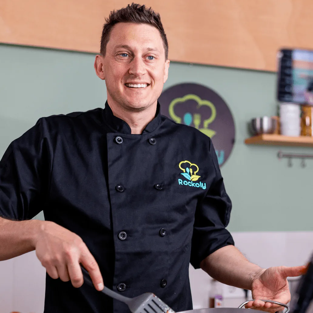 Smiling chef in a black Rockoly chef coat gesturing while leading a class