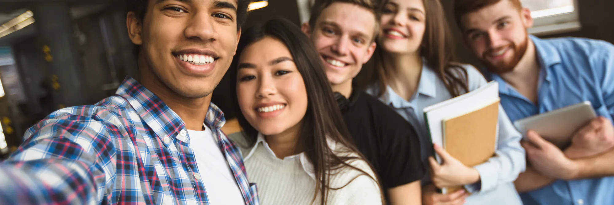 Five smiling young colleagues lean in for a group selfie holding notebooks and a tablet
