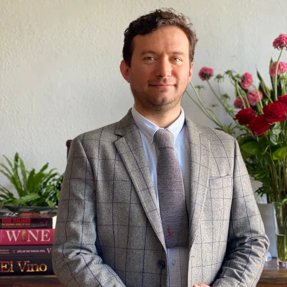 Sommelier Angelo Scimia in a grey checked blazer standing beside a shelf of wine books