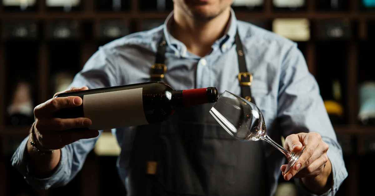 Sommelier in an apron tilting a bottle of red wine toward an empty glass in a wine cellar