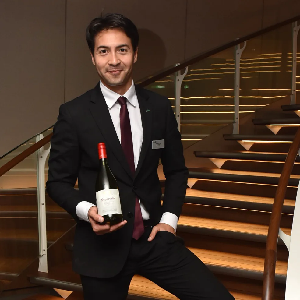 Sommelier Sebastian Pacheco in a dark suit holding a bottle of wine on a curved lit staircase