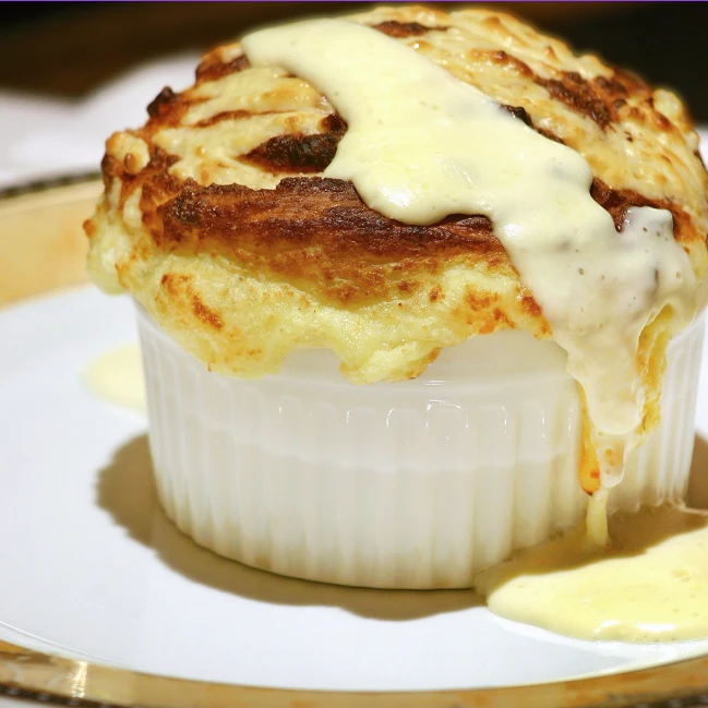Golden baked soufflé rising out of a white fluted porcelain ramekin, with cream sauce poured over the top and pooling on the plate