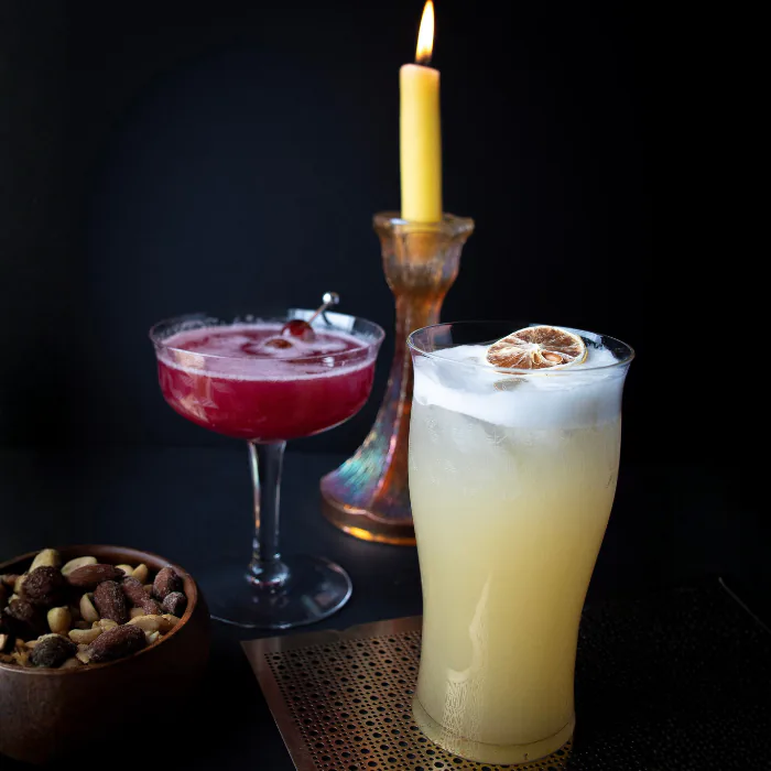 Ruby coupe cocktail and a tall foamy fizz with a dried lime wheel lit by a candle on a dark bar