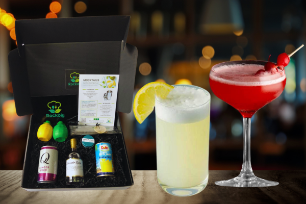 Cocktail kit box beside a frothy lemon gin fizz and a red coupe cocktail garnished with cherries