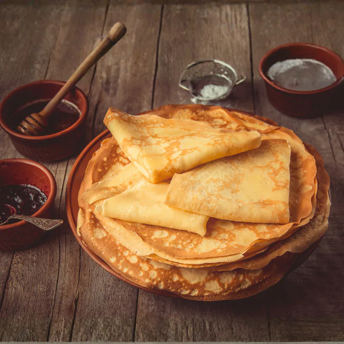 Stack of thin French crêpes, some folded, on a plate with honey, jam and cream in rustic bowls