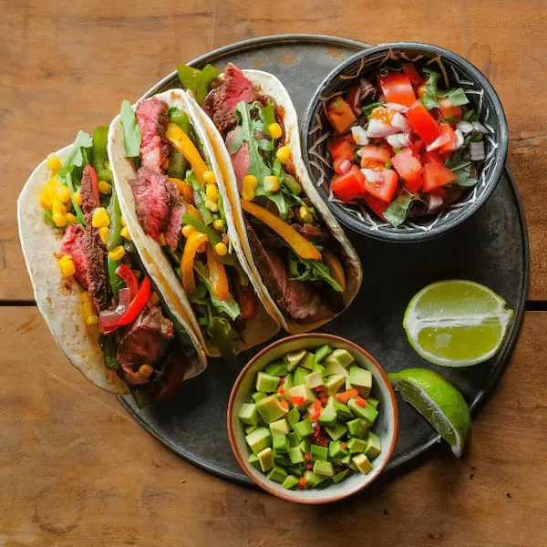 Three steak street tacos with peppers and corn, served with pico de gallo, avocado salsa and lime