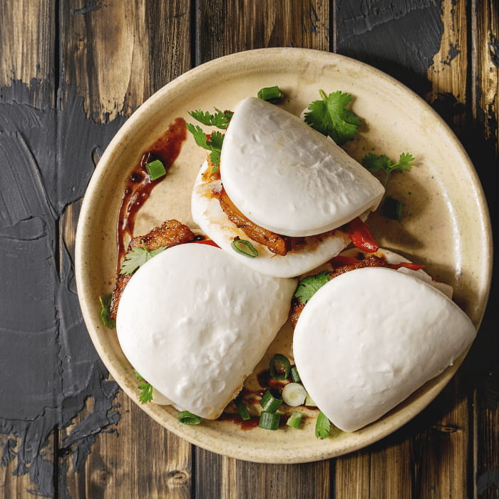 Three folded steamed bao buns with saucy filling, scallions and cilantro on a ceramic plate
