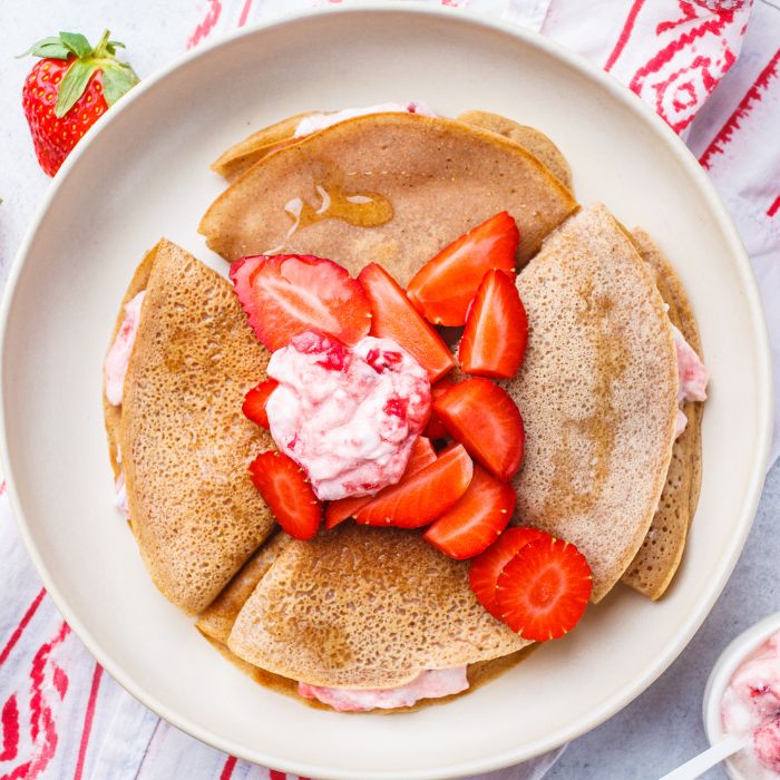Folded crepes on a plate topped with fresh strawberries and a spoonful of strawberry cream
