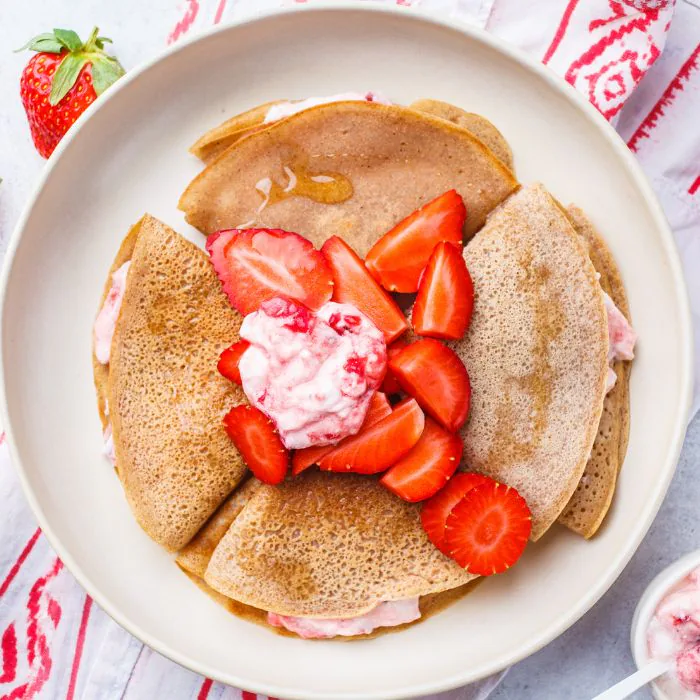 Folded crepes on a plate topped with fresh strawberries and a spoonful of strawberry cream