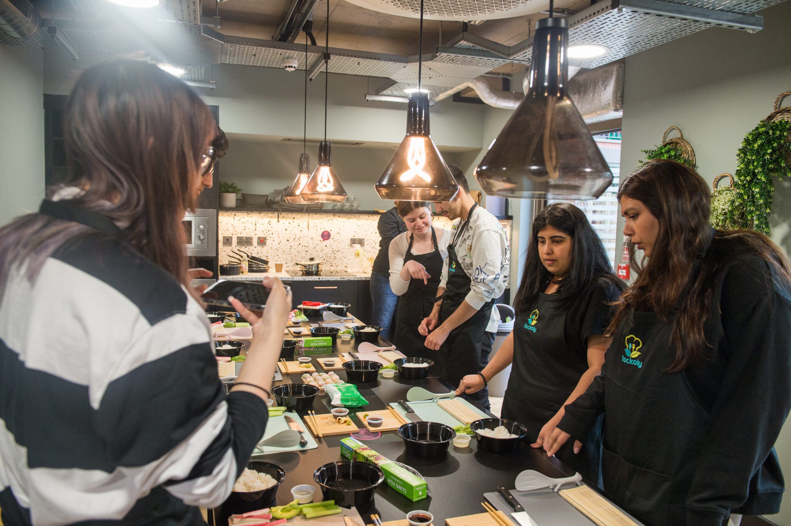 Group in aprons making sushi around a kitchen counter under copper pendant lamps