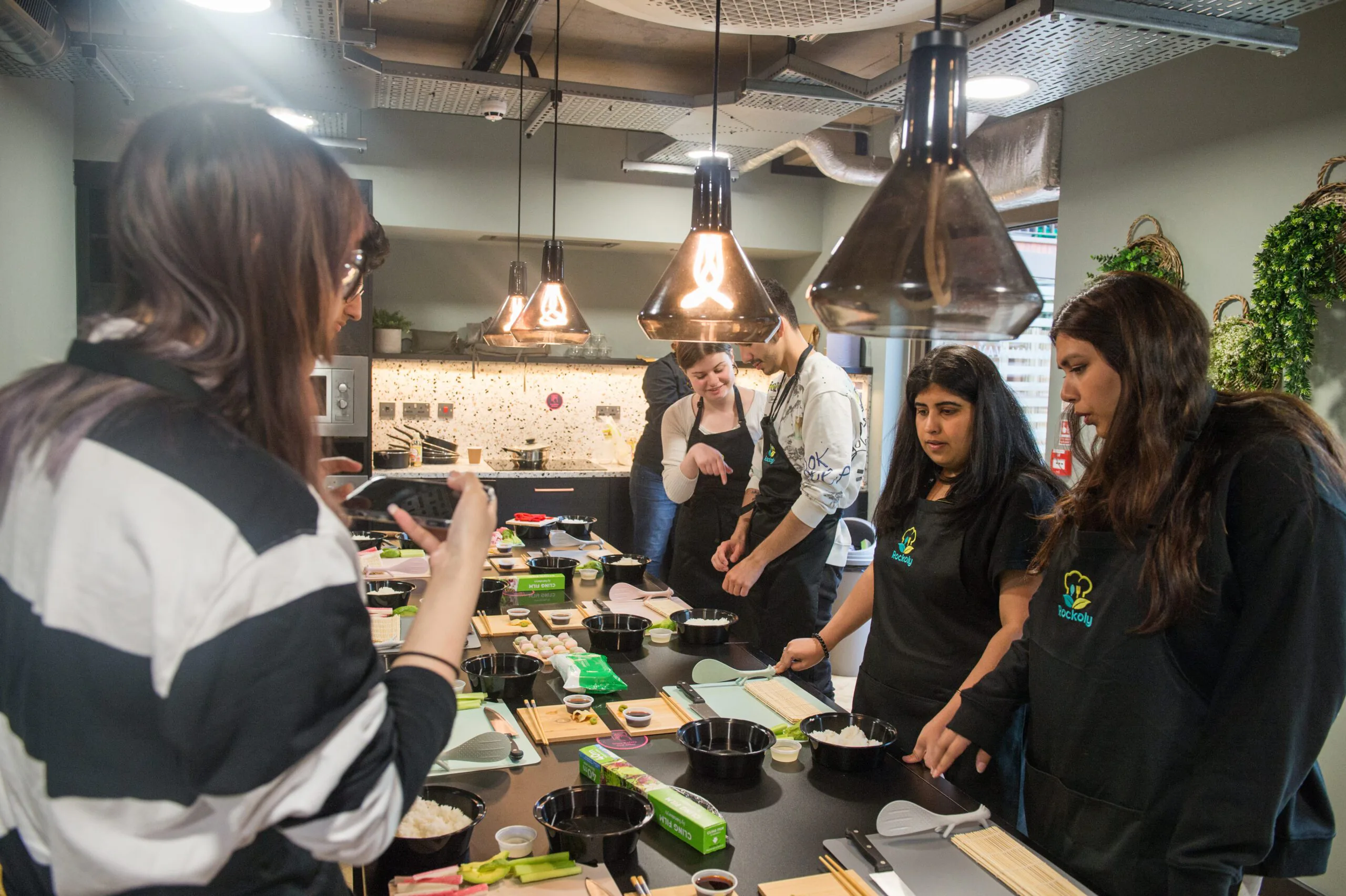 Group in aprons making sushi around a kitchen counter under copper pendant lamps
