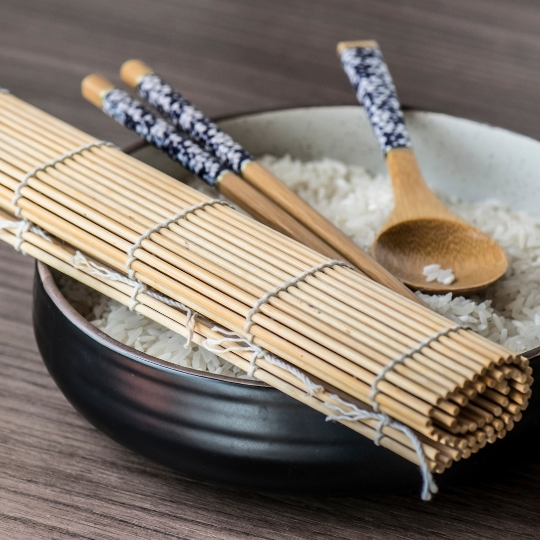 Bamboo sushi rolling mats, chopsticks and a wooden paddle resting on a bowl of sushi rice