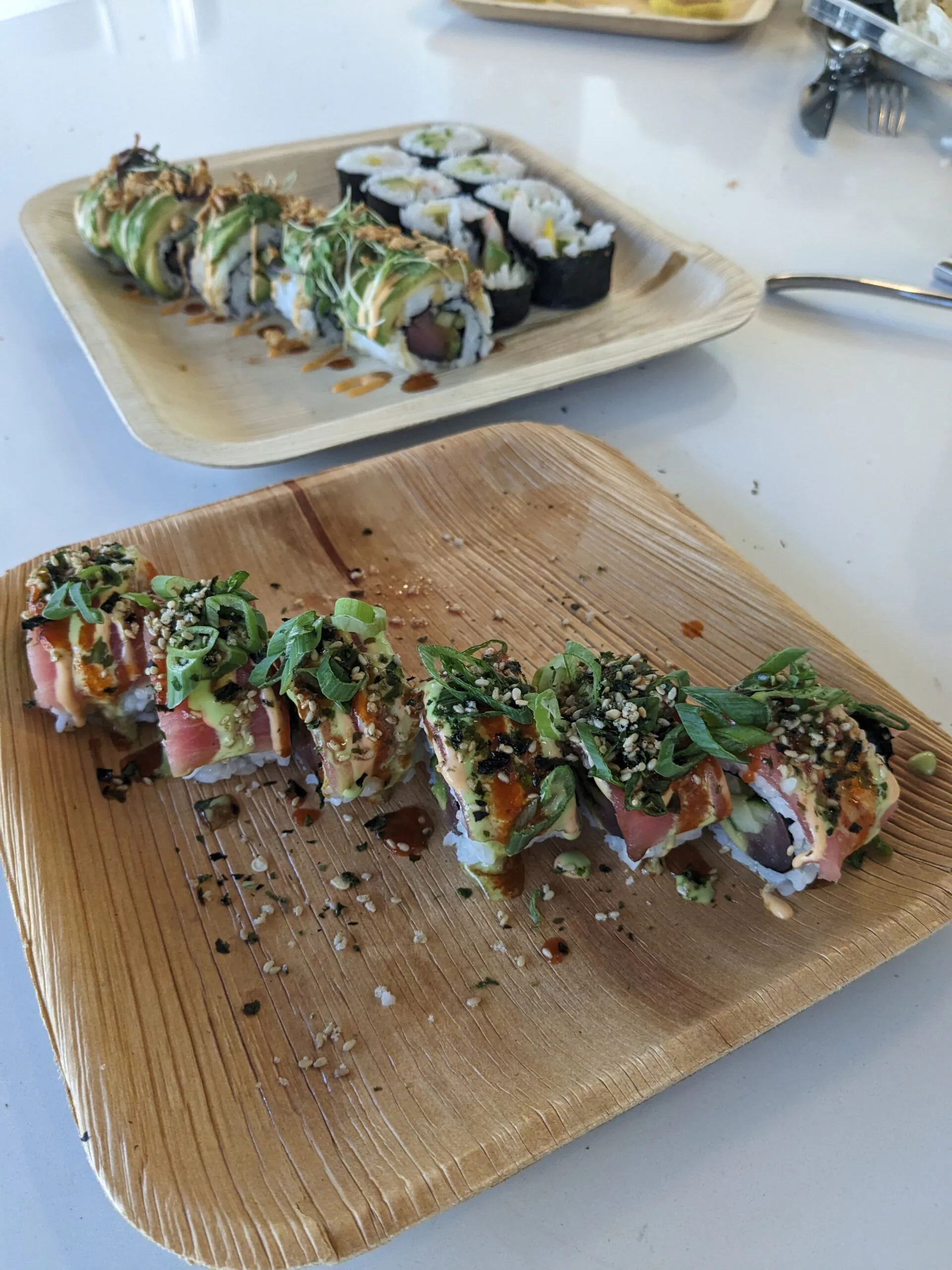 Two square palm-leaf plates of sushi rolls topped with avocado, scallions and sesame seeds