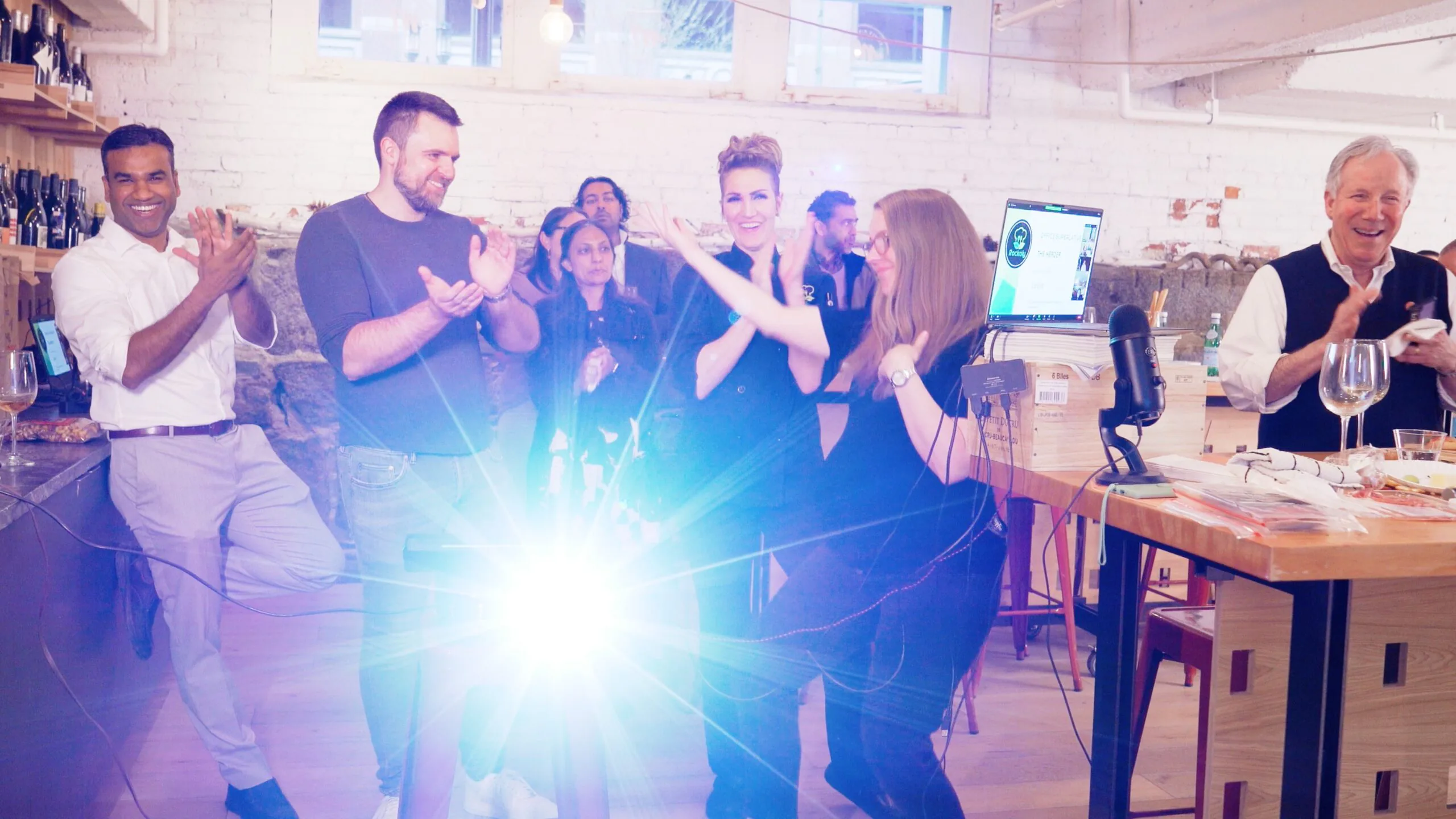 Guests clapping and cheering at a hybrid event, with remote colleagues on a screen and wine glasses nearby