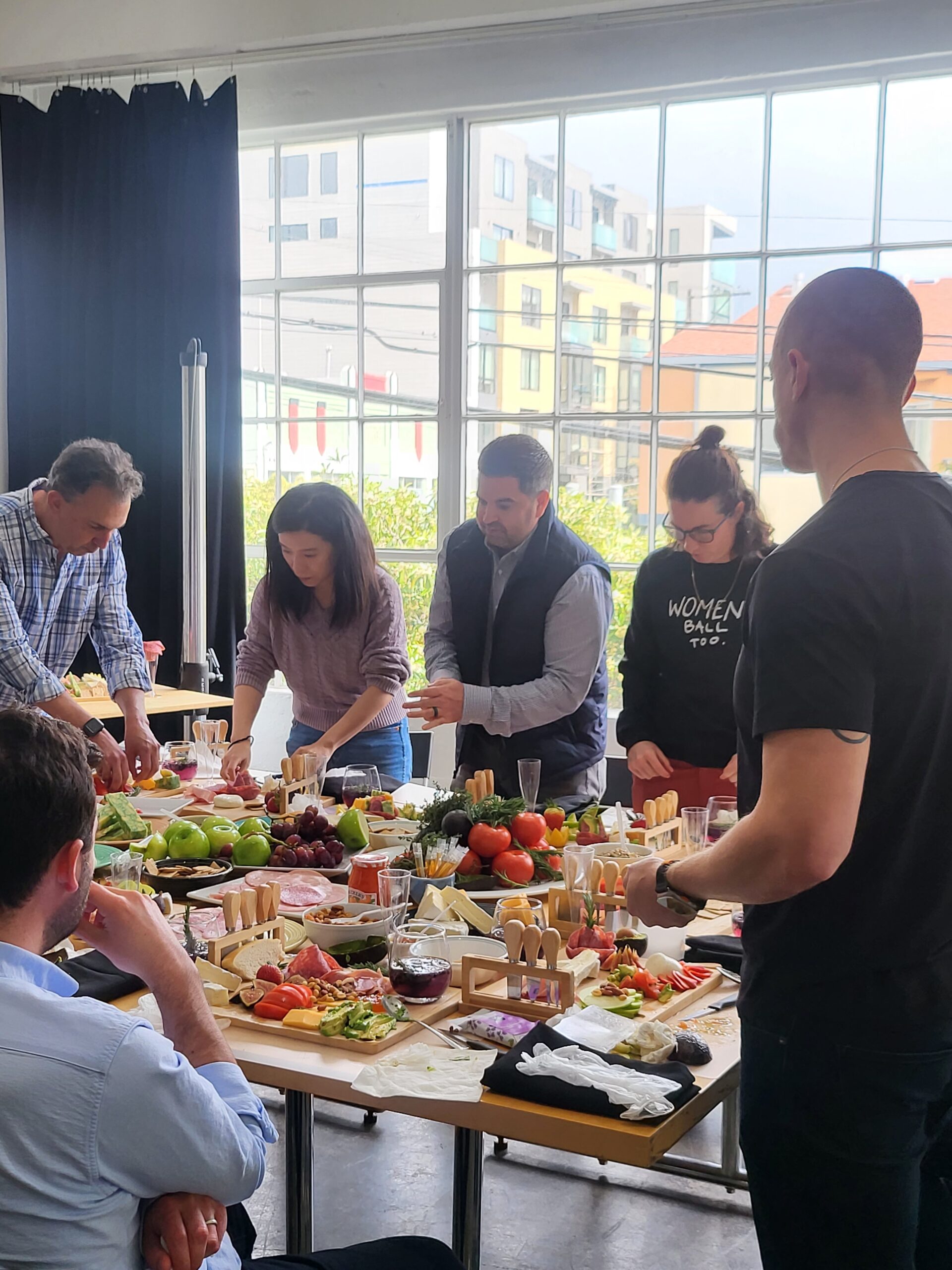 Colleagues gathered around a long grazing table of fruit, cheese and charcuterie by tall windows