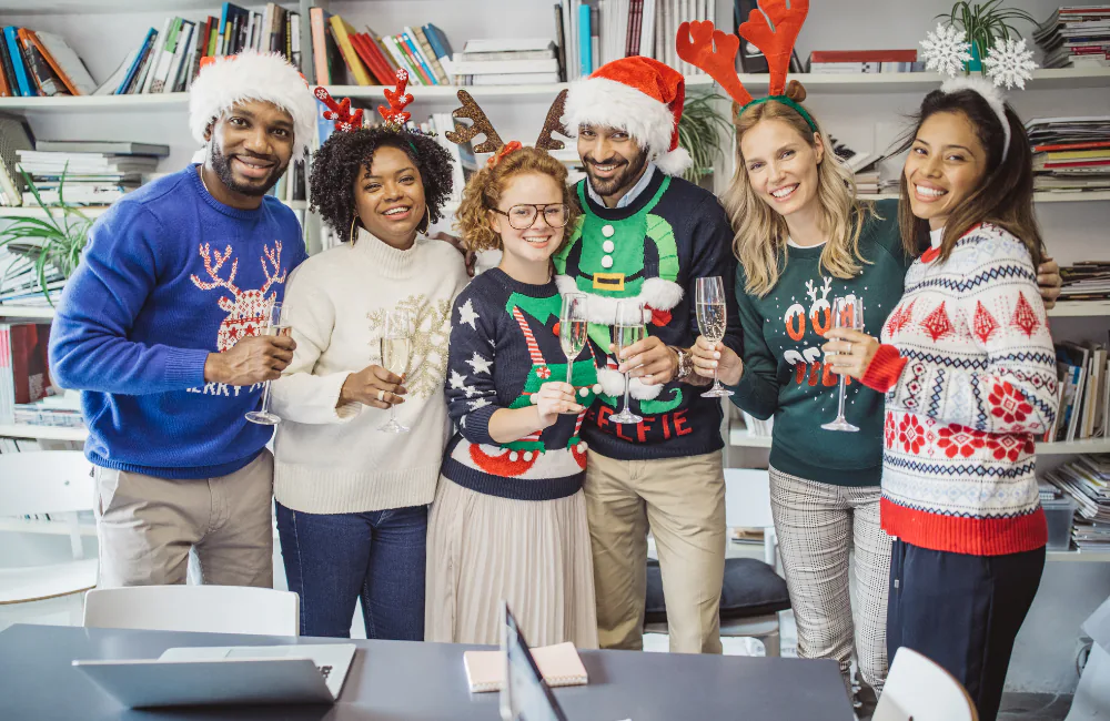 Six coworkers in holiday sweaters and Santa hats toasting with sparkling wine in an office
