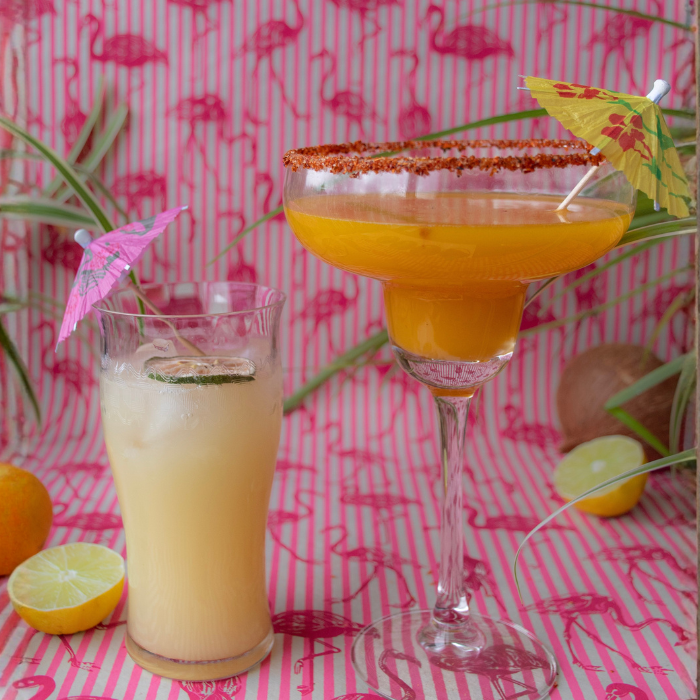 Two tropical drinks with paper umbrellas on flamingo-print fabric, one orange margarita and one cooler