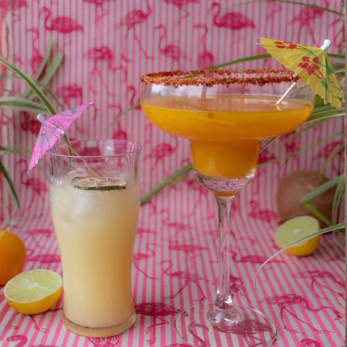 Two tropical drinks with paper umbrellas on flamingo-print fabric, one orange margarita and one cooler