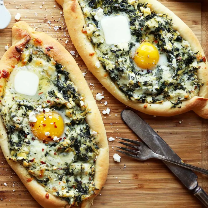 Two oval Turkish pide flatbreads topped with spinach, cheese and a runny egg yolk on a wooden board