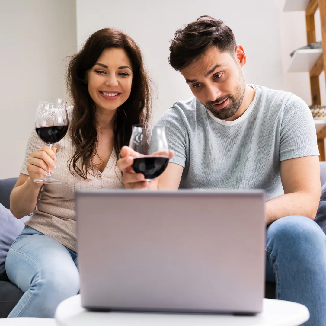 Two people on a sofa holding glasses of red wine while watching a laptop screen