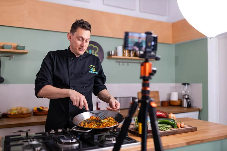 Chef in a black jacket tossing food in a pan while a phone on a tripod films him in a home kitchen