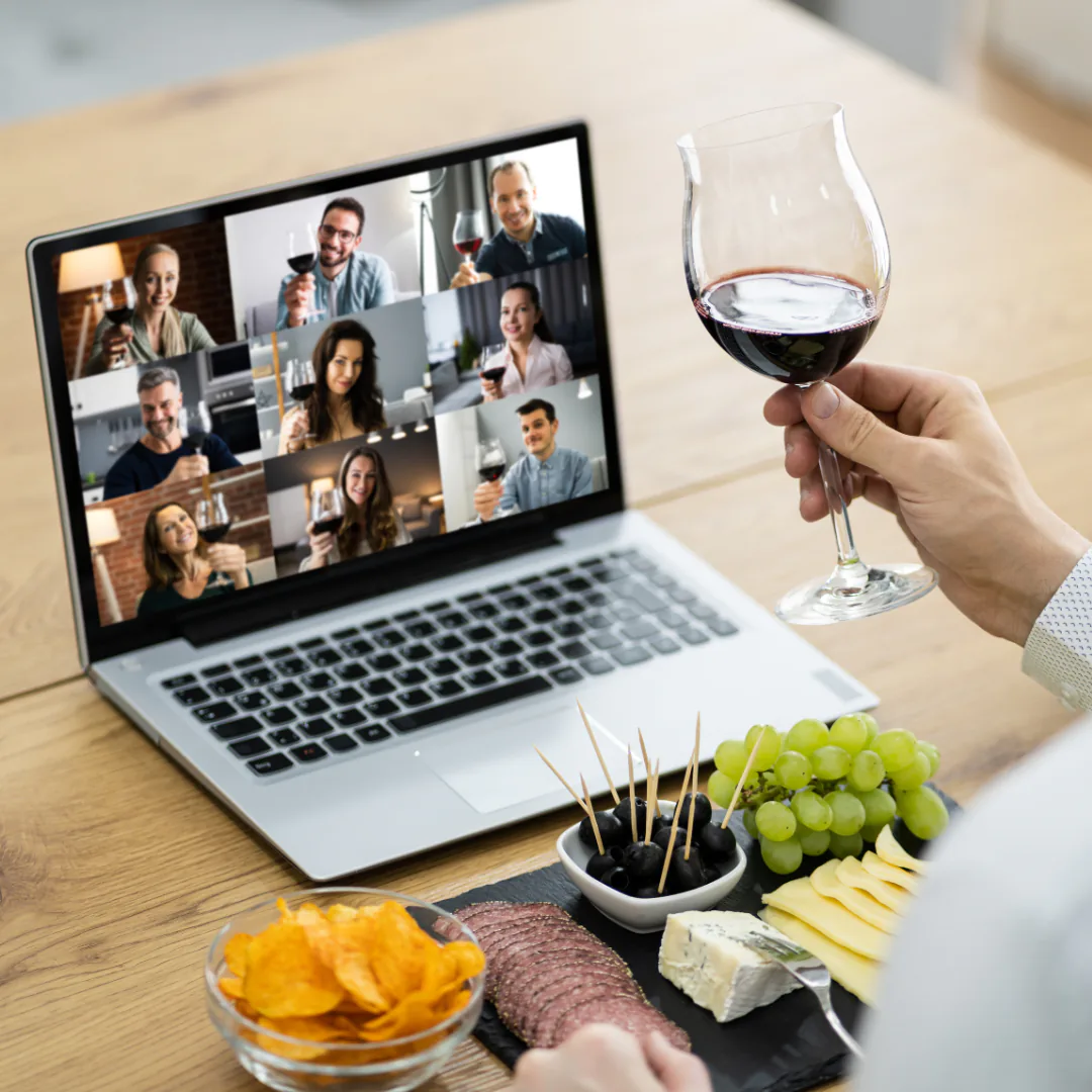 Hand raising a glass of red wine at a laptop video call while others toast, beside a cheese board