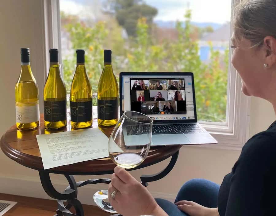 Woman holding a glass of white wine beside four bottles while participants appear on her laptop video call