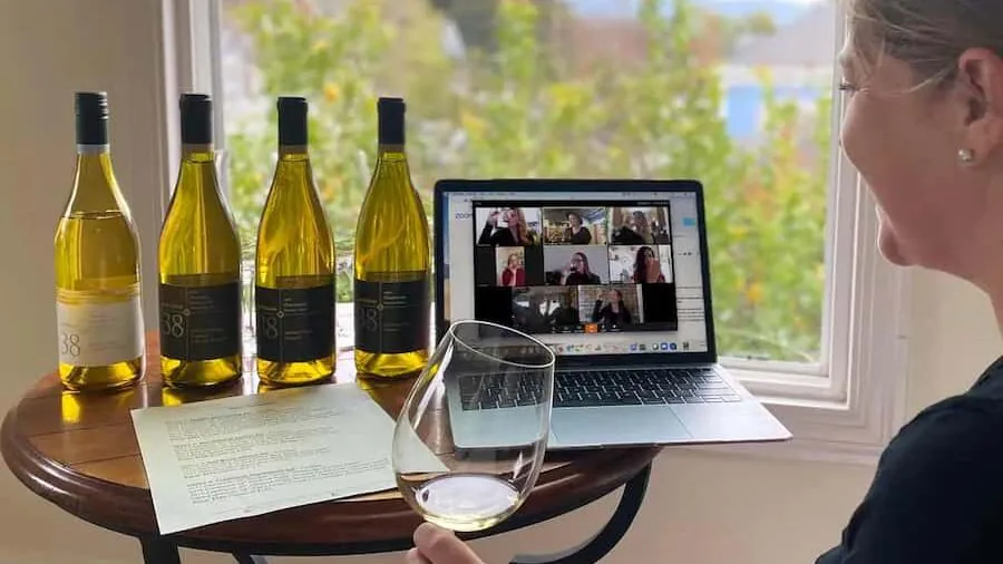 Woman holding a glass of white wine beside four bottles and a laptop showing a video-call grid
