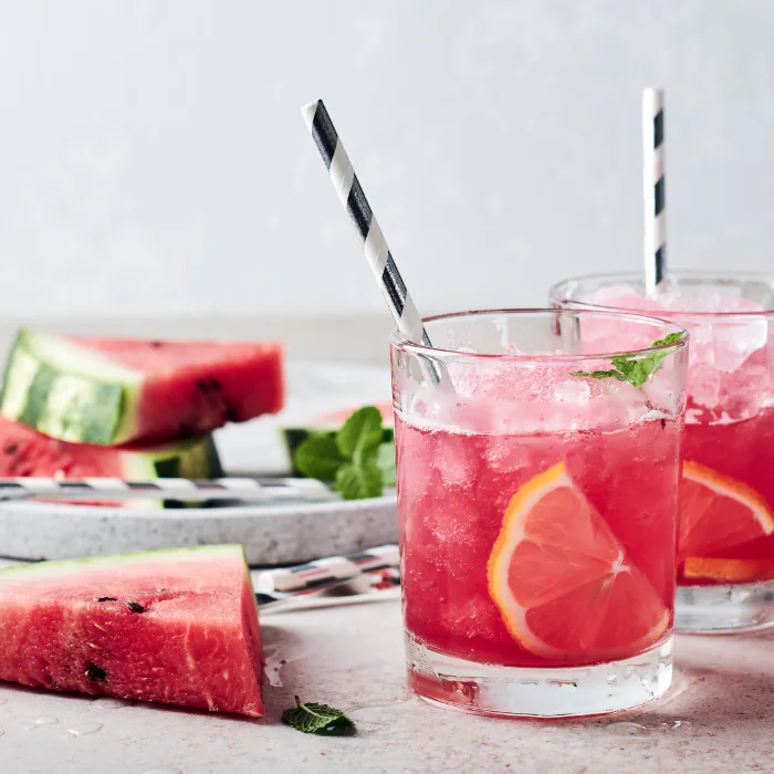 Two iced watermelon cocktails with grapefruit, mint and striped paper straws beside melon slices