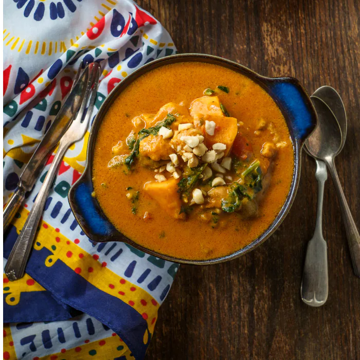 Overhead bowl of orange West African peanut stew garnished with herbs on a colorful patterned cloth