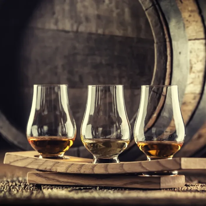Three Glencairn glasses of whiskey on a wooden flight board in front of an oak barrel