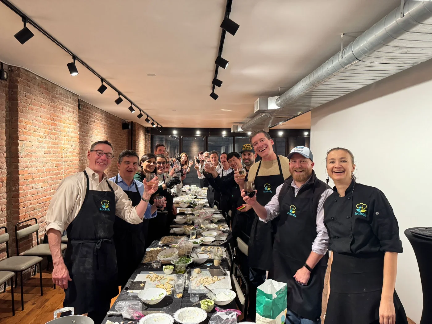 A team in Rockoly aprons raising glasses along a long table in a brick-walled kitchen, with their chef
