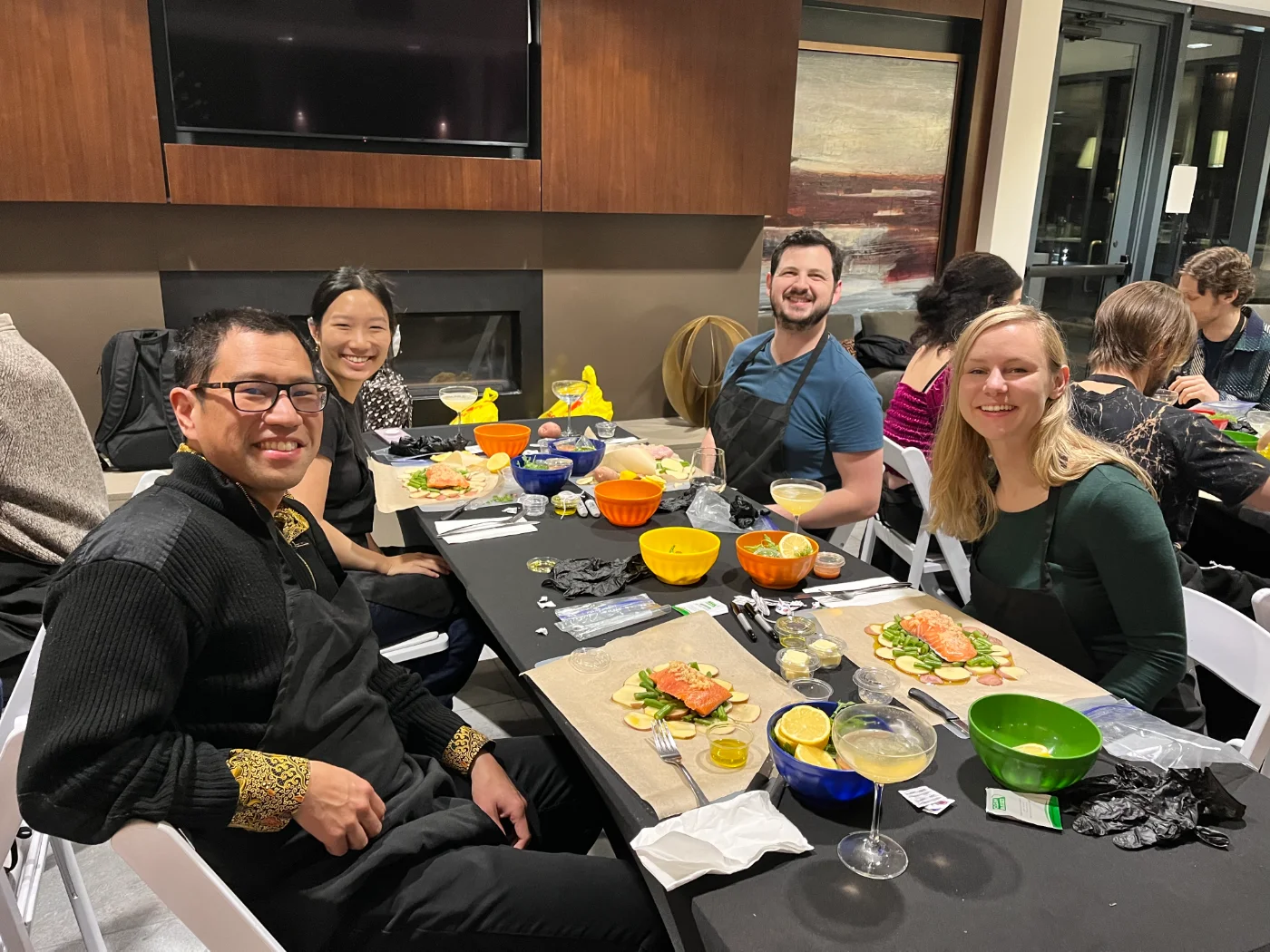 Guests smiling around a long table set with salmon en papillote, cocktails and coloured mixing bowls